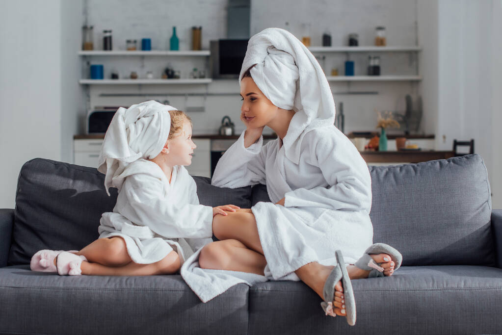 niño y mamá hablando mientras están sentados en el sofá con albornoces y toallas en la cabeza - Foto, Imagen