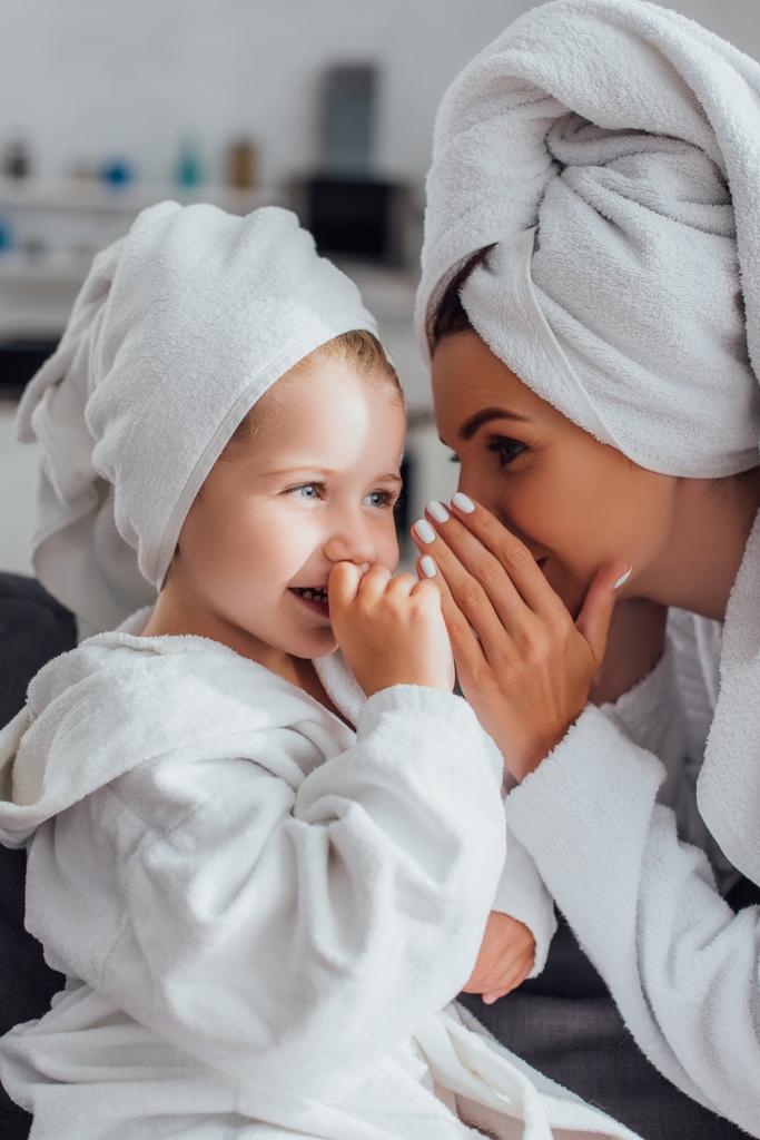 young mother covering mouth while telling secret to daughter while sitting together in bathrobes and towels on head - Photo, Image