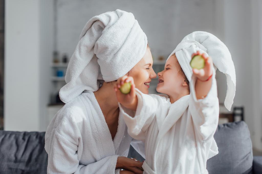 mujer joven con hija en batas blancas y toallas en la cabeza sentada cara a cara mientras la niña sostiene rodajas de pepino fresco - Foto, Imagen