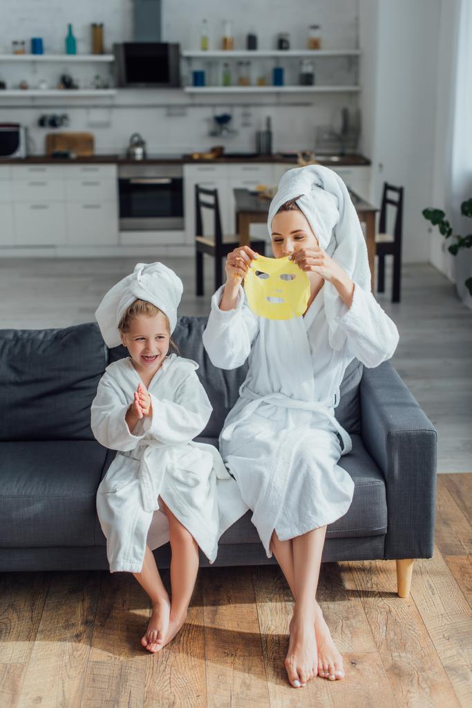 young woman holding face mask while sitting on sofa with daughter in bathrobes and towels on heads - Photo, Image