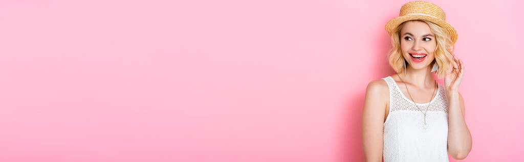 horizontale Ernte von Frau mit Strohhut berührt Haare und schaut weg auf rosa  - Foto, Bild