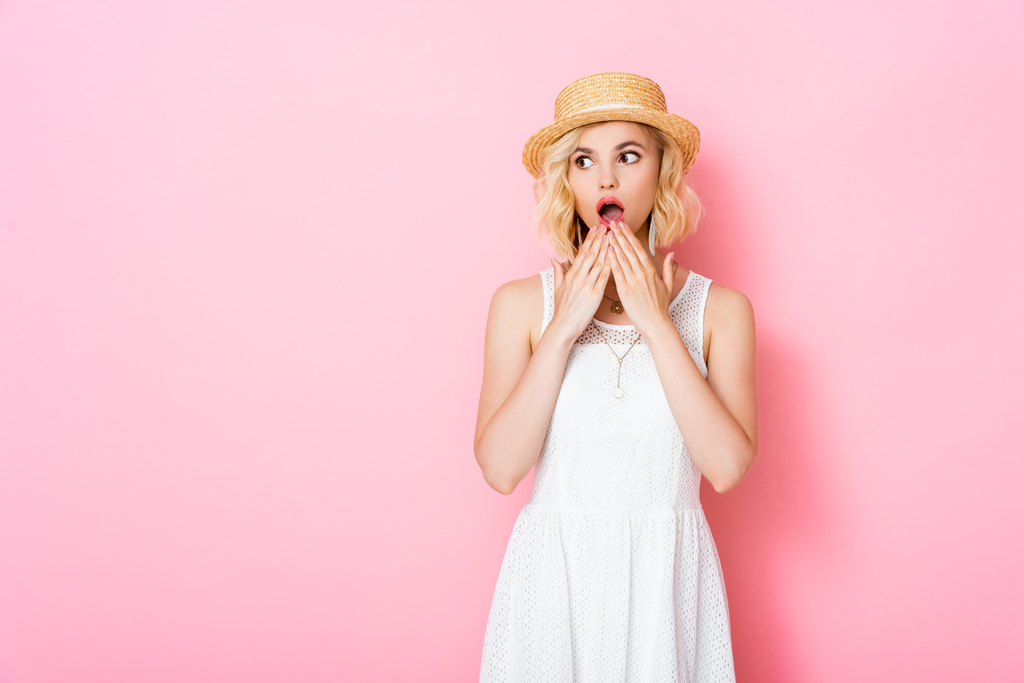 шокированная молодая женщина в соломенной шляпе, покрывающей рот и смотрящая на розовый  - Фото, изображение