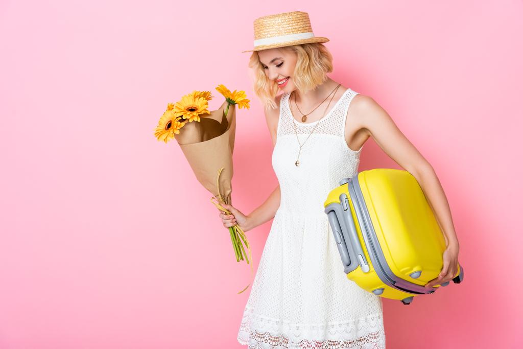 młoda kobieta w słomkowym kapeluszu z żółtymi kwiatami i bagażem na różowym  - Zdjęcie, obraz
