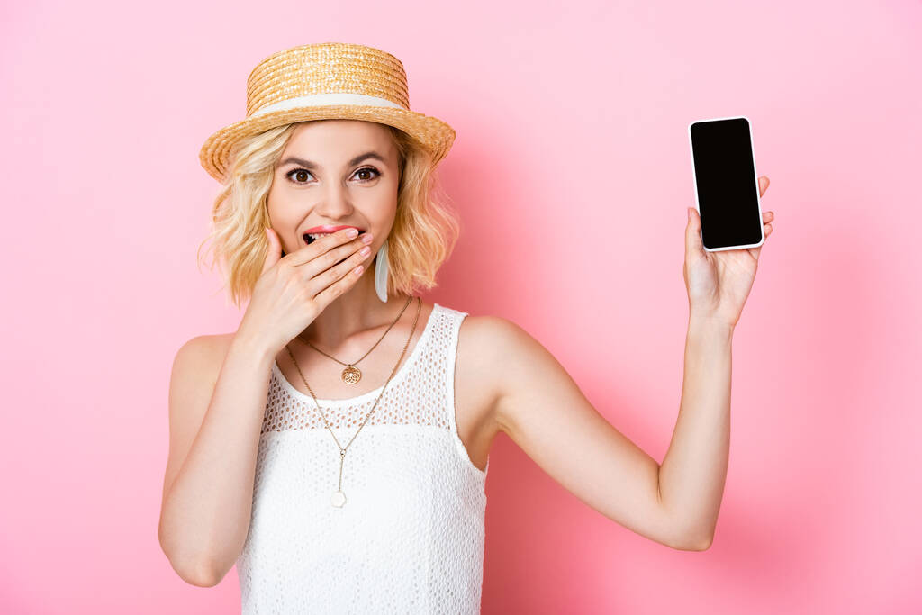 Frau mit Strohhut hält Smartphone mit leerem Bildschirm und verdeckt Mund auf rosa  - Foto, Bild
