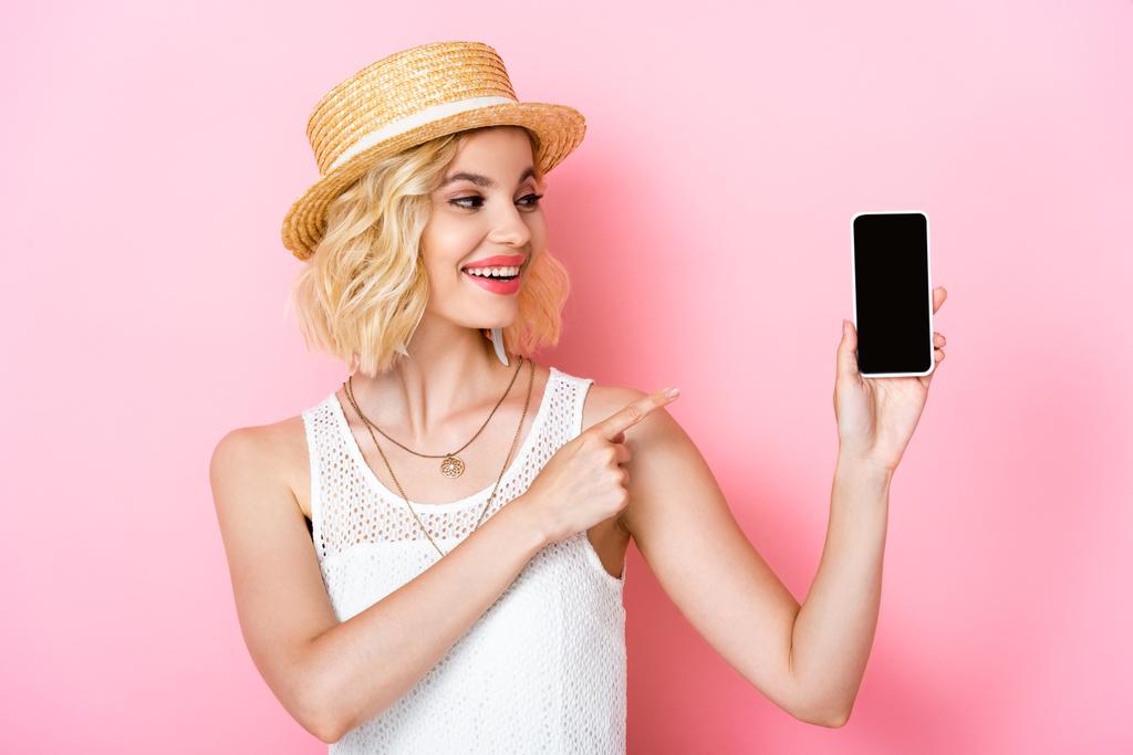 женщина в соломенной шляпе указывает пальцем на смартфон с пустым экраном на розовом  - Фото, изображение