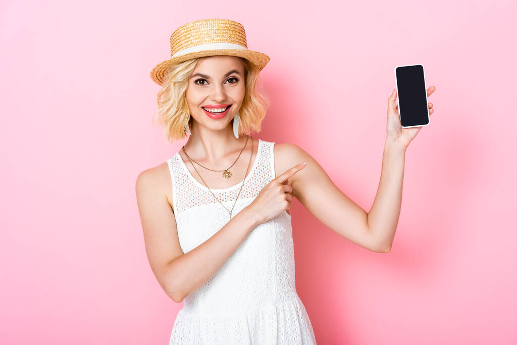 kobieta w słomkowym kapeluszu wskazując palcem na smartfona z pustym ekranem na różowym  - Zdjęcie, obraz