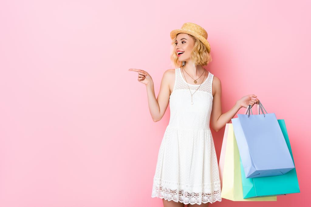 Frau mit Strohhut zeigt mit Finger und hält Einkaufstüten auf rosa  - Foto, Bild