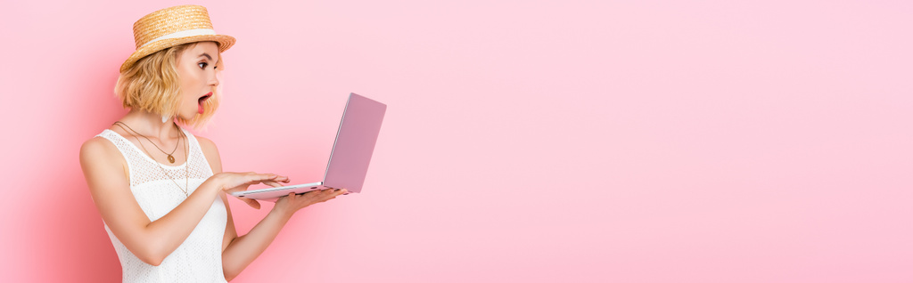 panoramische oriëntatie van geschokte vrouw in stro hoed op zoek naar laptop op roze - Foto, afbeelding