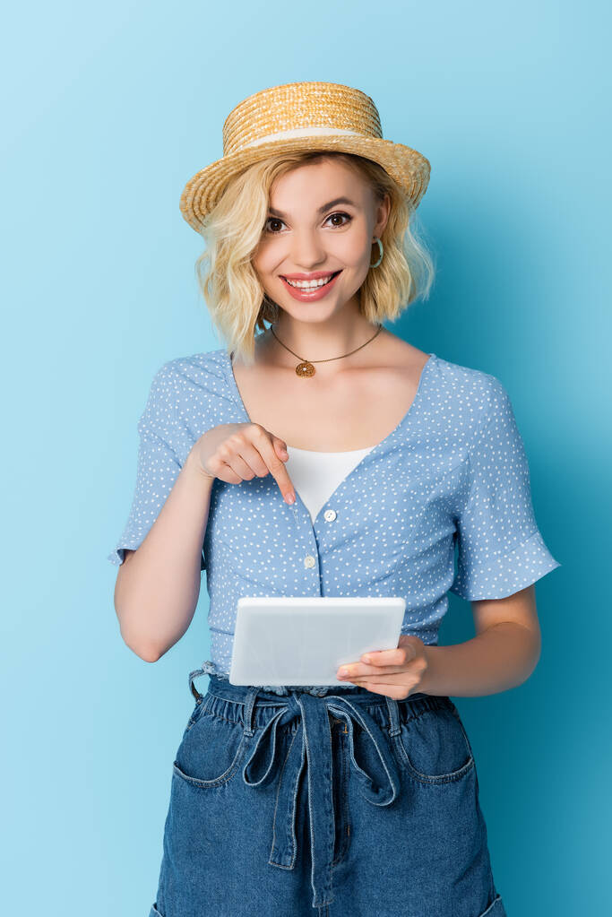 junge Frau mit Strohhut zeigt mit Finger auf digitales Tablet auf blau  - Foto, Bild
