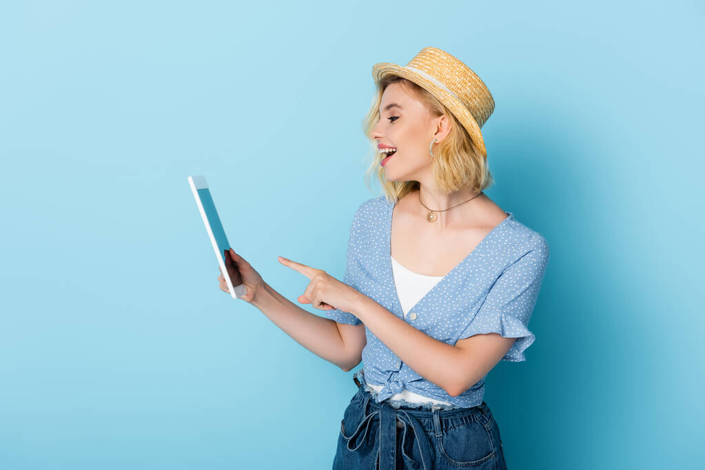 femme excitée en chapeau de paille pointant avec le doigt à la tablette numérique sur bleu  - Photo, image