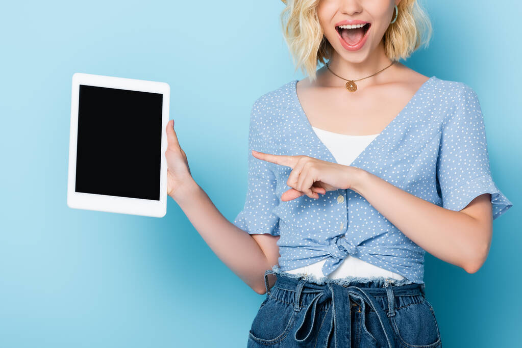 Ausgeschnittene Ansicht einer aufgeregten Frau, die mit dem Finger auf ein digitales Tablet mit leerem Bildschirm auf blauem Hintergrund zeigt  - Foto, Bild