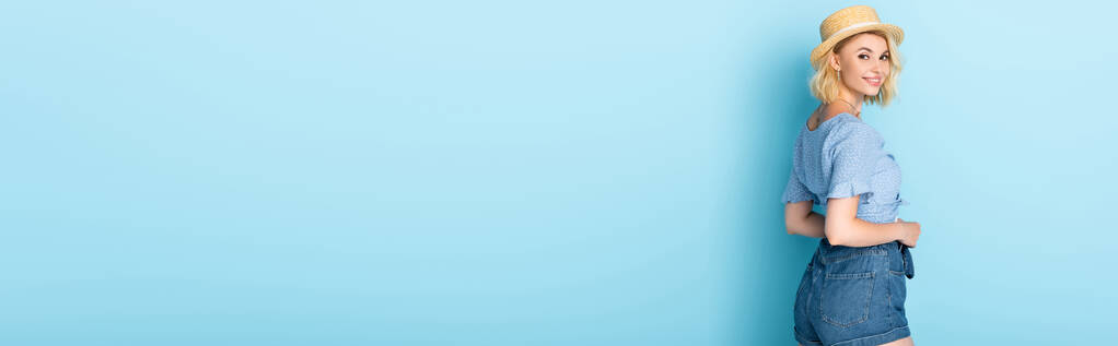 cultivo panorámico de mujer joven en pantalones cortos y sombrero de paja de pie en azul - Foto, Imagen