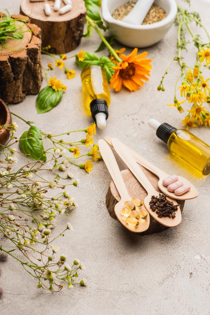 selective focus of wildflowers, herbs, bottles and pills in wooden spoons on concrete background, naturopathy concept - Photo, Image