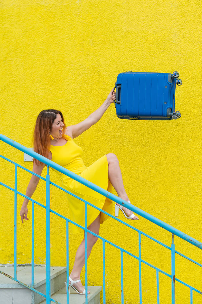 Μια χαρούμενη κοκκινομάλλα με κίτρινο φόρεμα κατεβαίνει τις σκάλες και κουνάει με χαρά μια μπλε βαλίτσα. Το κορίτσι ετοιμάζεται για το ταξίδι. Καλοκαιρινές διακοπές. - Φωτογραφία, εικόνα