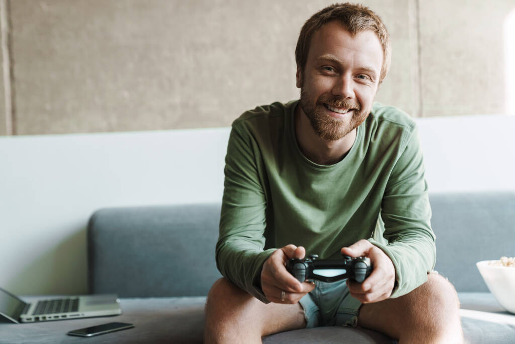 Foto de alegre pelirrojo jugando videojuego con joystick mientras está sentado en el sofá en casa - Foto, imagen