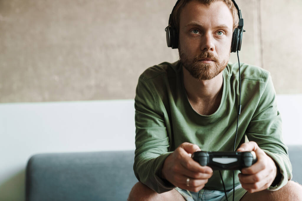 Kuva keskittynyt mies yllään kuulokkeet pelaa videopeliä joystick istuessaan sohvalla kotona - Valokuva, kuva