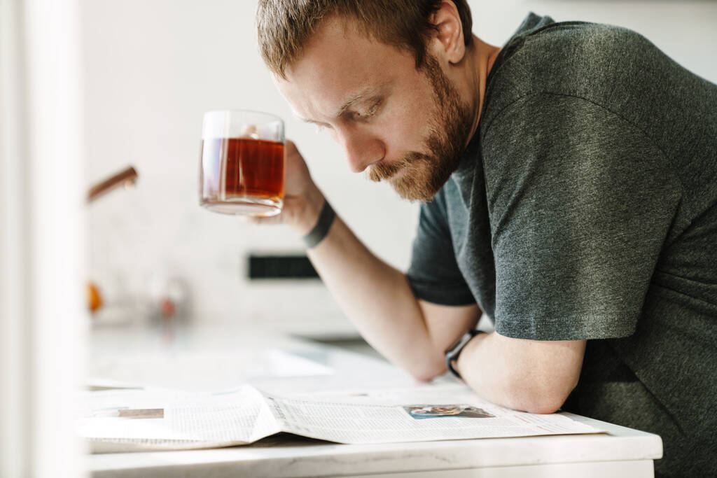 Фото серьезного рыжего человека с бородой, читающего газету во время чая на кухне дома - Фото, изображение