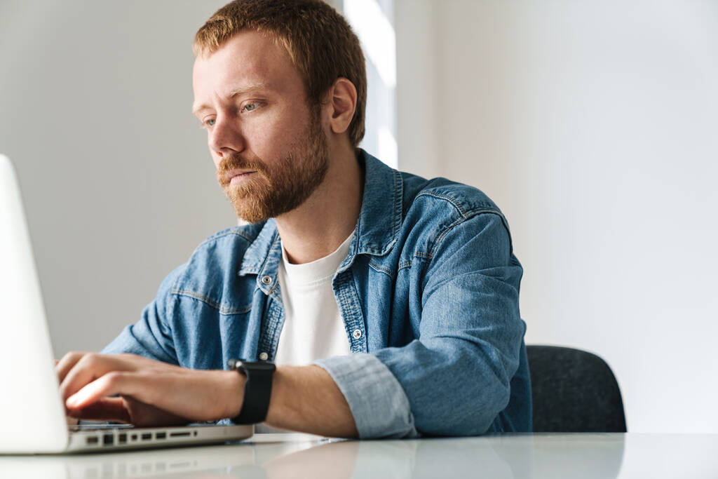 Photo d'un bel homme avec une barbe rouge travaillant sur un ordinateur portable assis à la table à l'intérieur - Photo, image