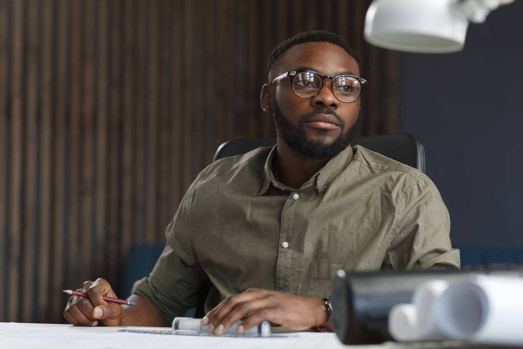 Αφρο-Αμερικανός αρχιτέκτονας που εργάζεται στο γραφείο με τα σχέδια. Μηχανικός επιθεωρήσει αρχιτεκτονικό σχέδιο, σχεδιάζοντας ένα κατασκευαστικό έργο. Πορτρέτο του μαύρου όμορφου άντρα που κάθεται στο χώρο εργασίας. Επιχειρηματική έννοια - Φωτογραφία, εικόνα