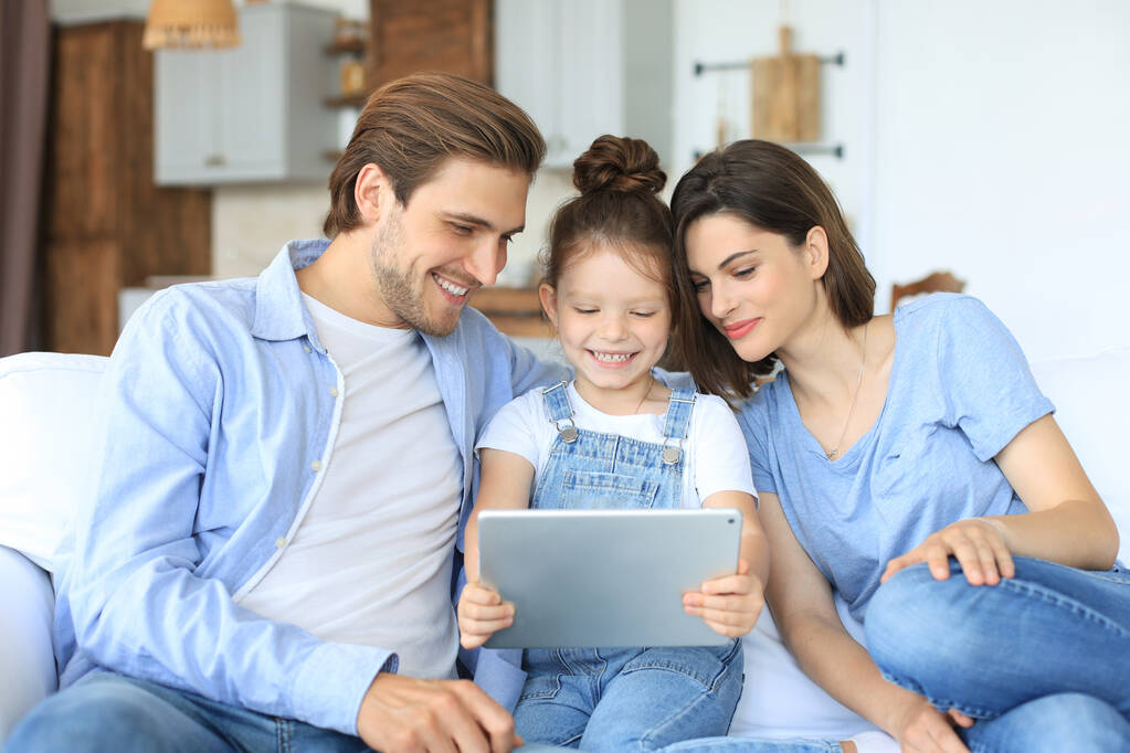 Padres jóvenes amistosos positivos con una hijita sonriente sentada en el sofá juntos respondiendo a una videollamada en una tableta digital mientras se relaja en casa el fin de semana - Foto, imagen