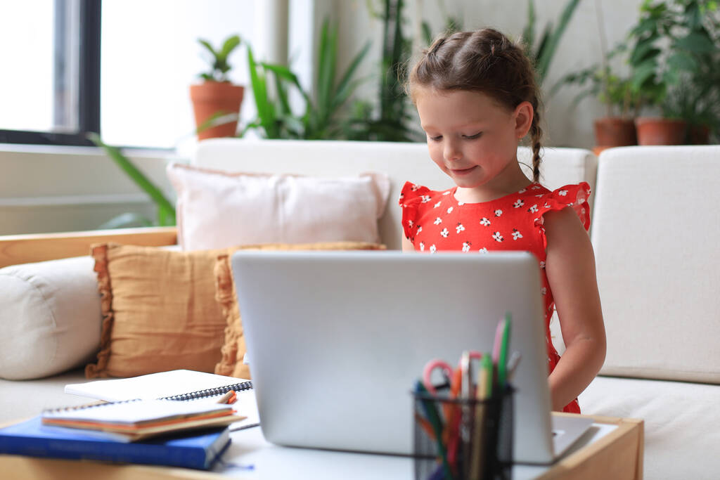 Дистанционное обучение. Веселая маленькая девочка с помощью ноутбука компьютер изучает через систему электронного обучения онлайн - Фото, изображение