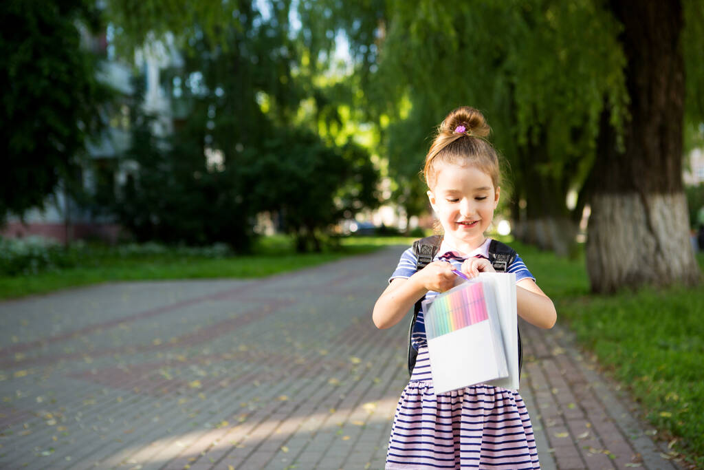 Uma menina de aparência caucasiana em um uniforme escolar com uma mochila e o livro e um conjunto de marcadores. de volta à escola. Escola primária, desenvolvendo atividades para pré-escolares. Espaço para texto
 - Foto, Imagem