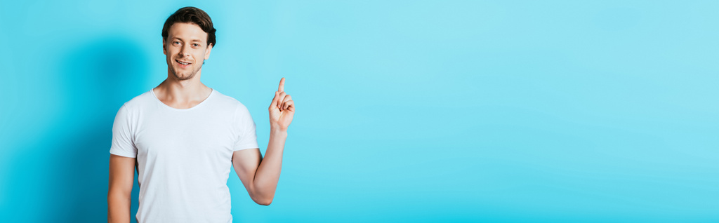 Panoramic shot of man in white t-shirt pointing with finger on blue background - Photo, Image