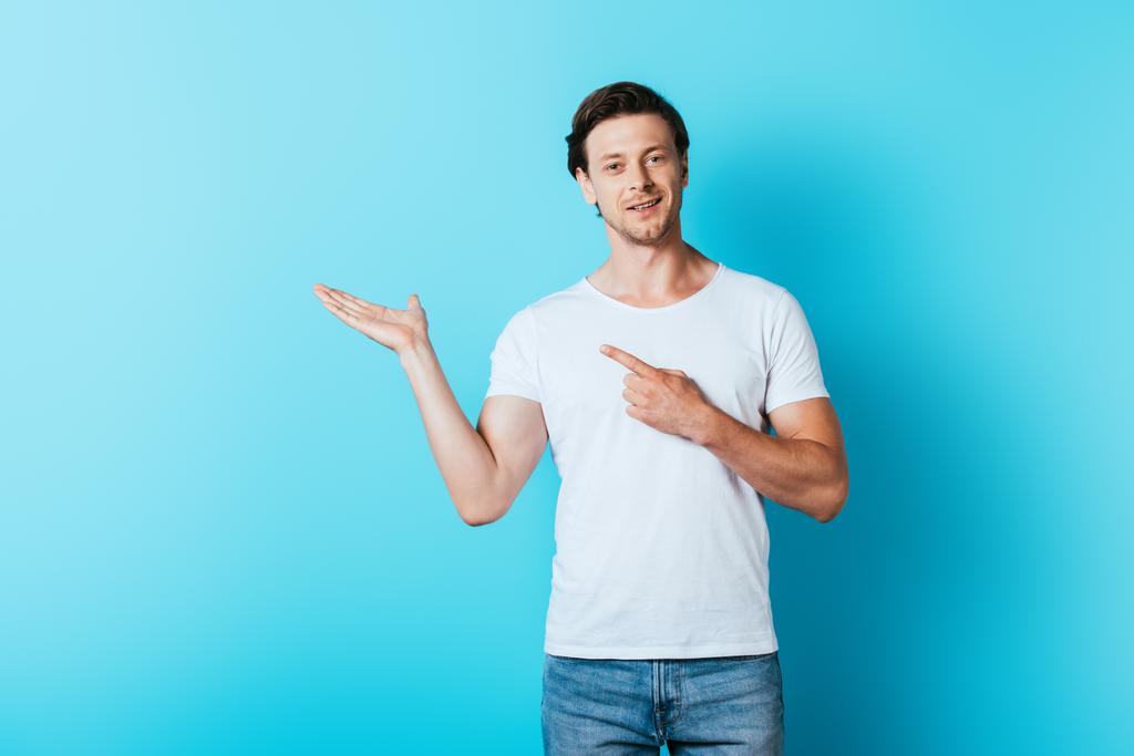 Man in white t-shirt pointing with hand and finger on blue background - Photo, Image