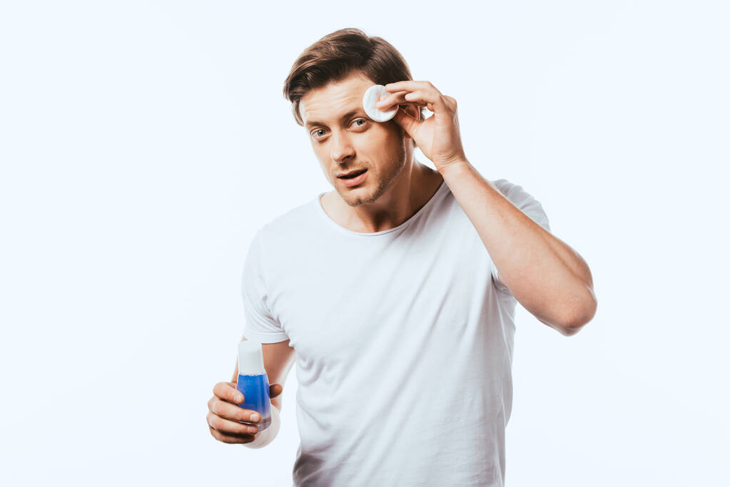 Giovane uomo guardando la fotocamera mentre si applica lozione con batuffolo di cotone isolato su bianco - Foto, immagini