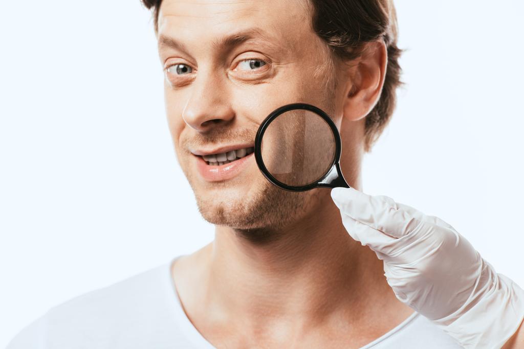 Dermatologo tenendo lente d'ingrandimento vicino guancia del paziente isolato su bianco - Foto, immagini