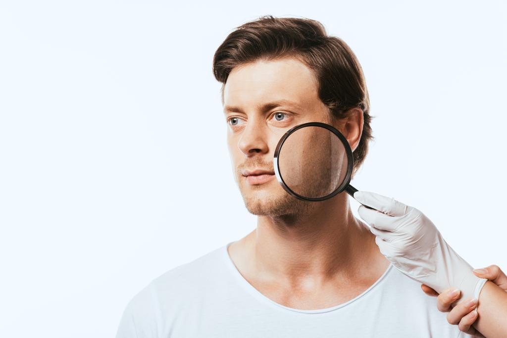 Mladý muž dívá pryč, zatímco dermatolog v latexové rukavici drží loupe izolované na bílém - Fotografie, Obrázek