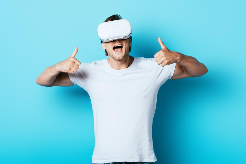 Νεαρός άνδρας με ακουστικά VR δείχνει τους αντίχειρες επάνω στο μπλε φόντο - Φωτογραφία, εικόνα