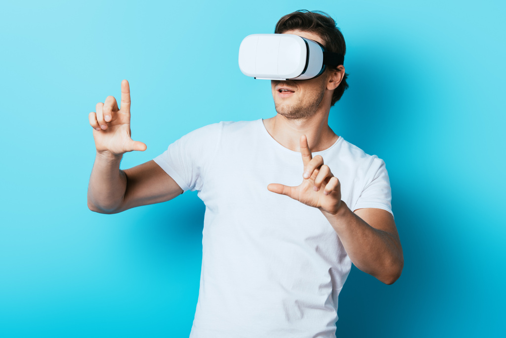 Junger Mann zeigt mit Fingern, während er Virtual-Reality-Headset auf blauem Hintergrund benutzt - Foto, Bild