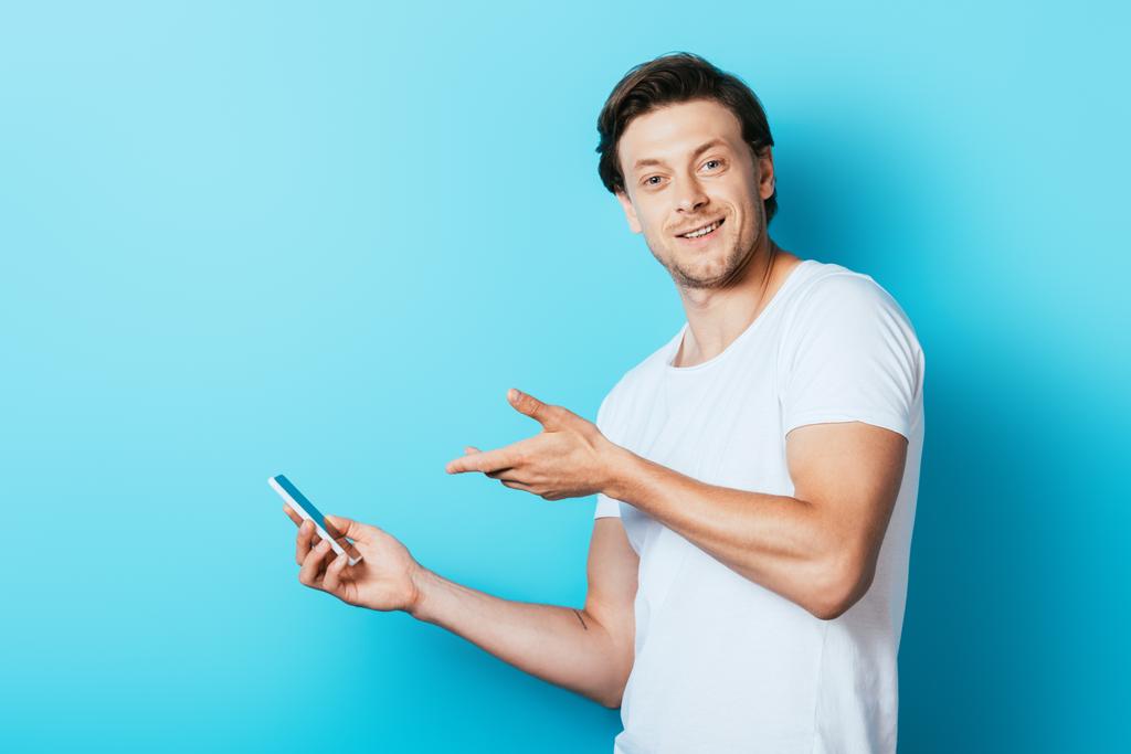 Junger Mann im weißen T-Shirt zeigt mit der Hand auf Smartphone auf blauem Hintergrund - Foto, Bild