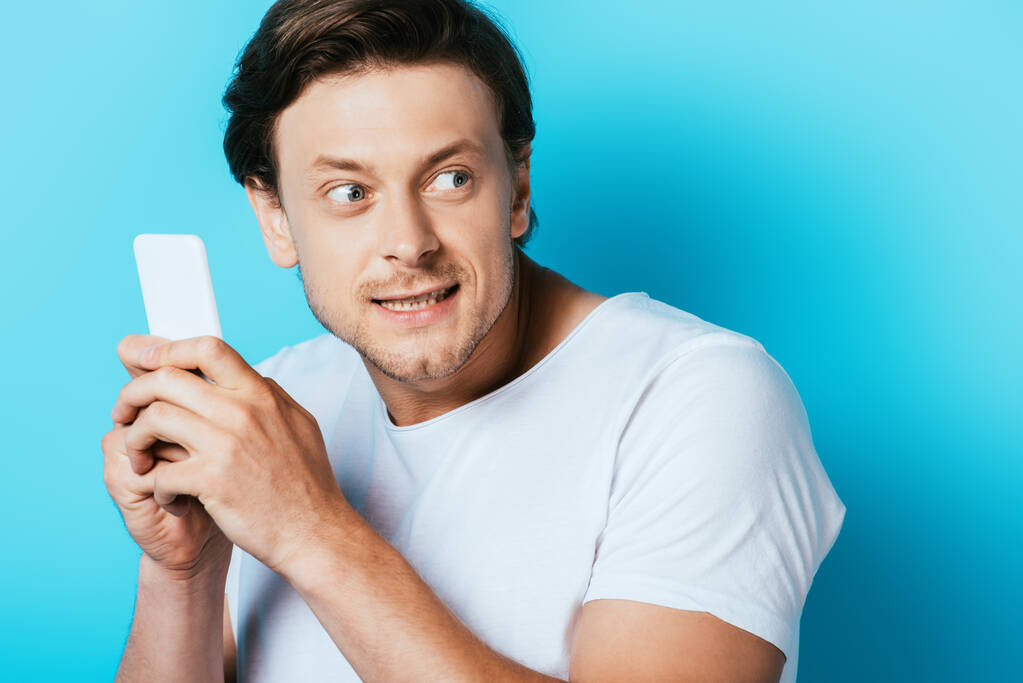 Tricky άνθρωπος κρατώντας smartphone και κοιτάζοντας μακριά σε μπλε φόντο - Φωτογραφία, εικόνα