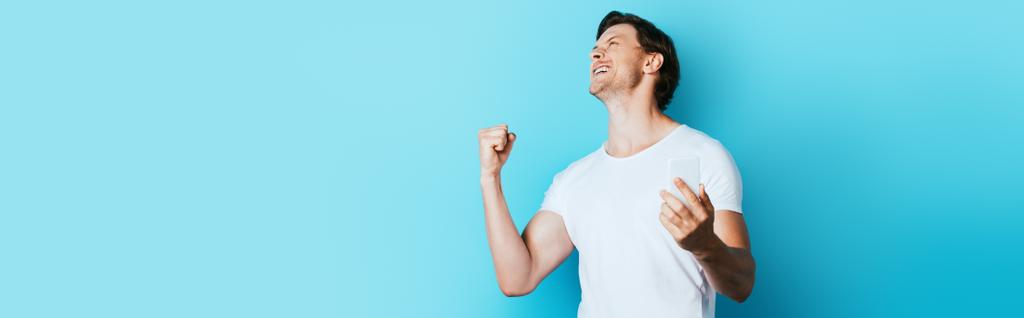 Panoramic shot of man showing yeah gesture and holding smartphone on blue background - Photo, Image