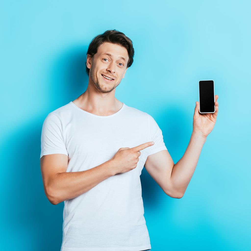 Junger Mann im weißen T-Shirt zeigt mit dem Finger auf Smartphone auf blauem Hintergrund - Foto, Bild