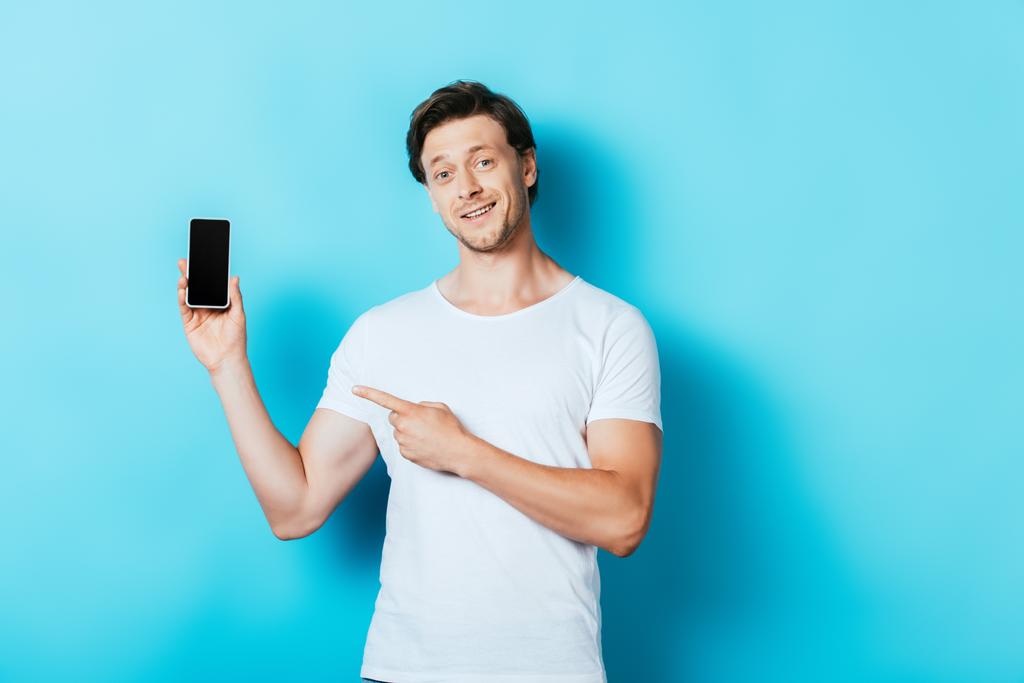 Man in white t-shirt pointing at smartphone on blue background - Photo, Image