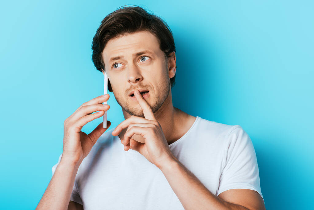 Задумчивый человек с пальцем возле губ разговаривает на смартфоне на синем фоне - Фото, изображение