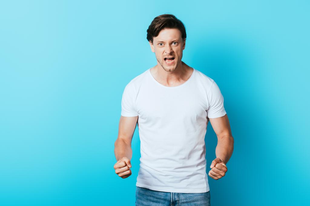 Irrated man in white t-shirt with hands in fists looking at camera on blue background - Photo, Image