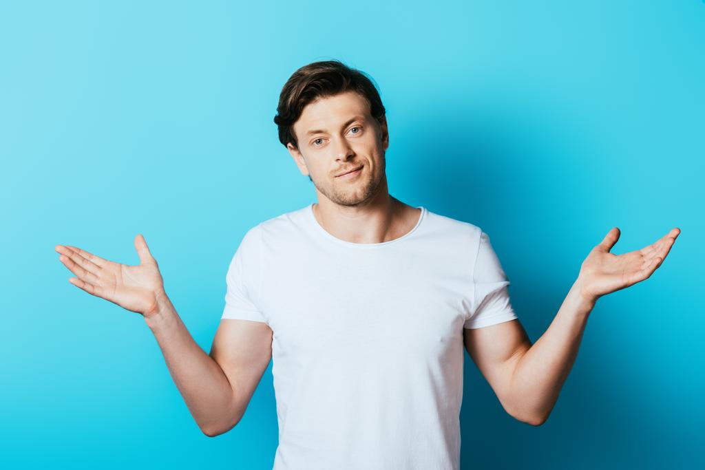 Muž v bílém tričku ukazuje pokrčení rameny gesto na modrém pozadí - Fotografie, Obrázek