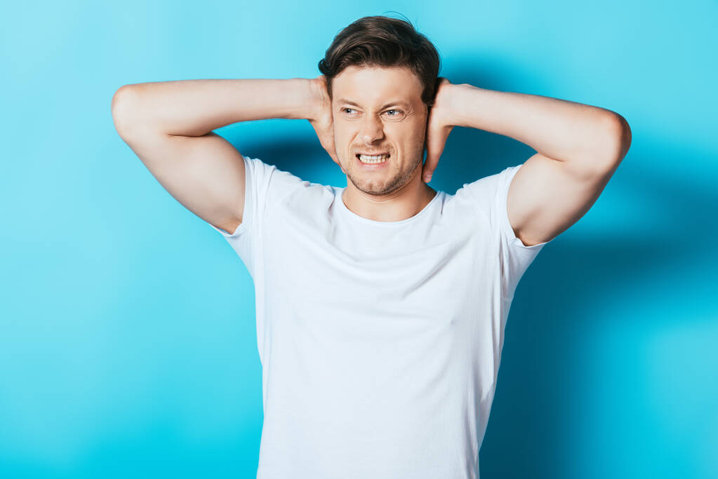 Angry man in white t-shirt covering ears with hands on blue background - Photo, Image