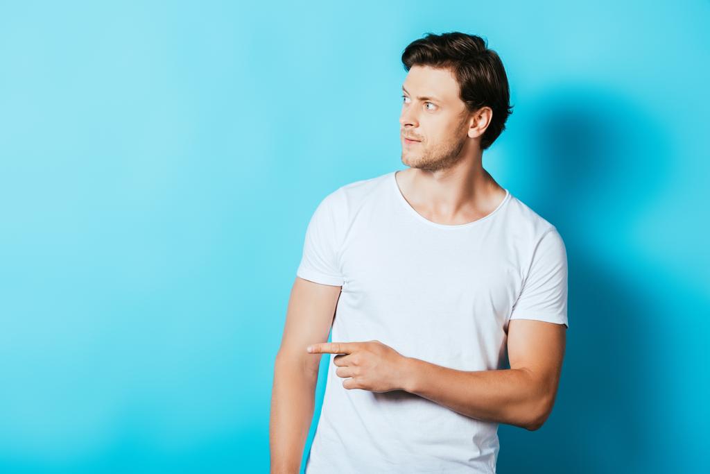 Hombre seguro en camiseta blanca apuntando con el dedo sobre fondo azul - Foto, Imagen