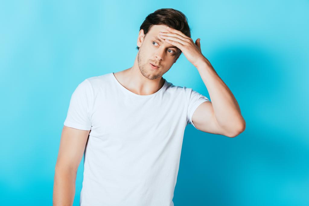 Νεαρός άνδρας με λευκό t-shirt αγγίζει το μέτωπο ενώ κοιτάζει μακριά στο μπλε φόντο - Φωτογραφία, εικόνα