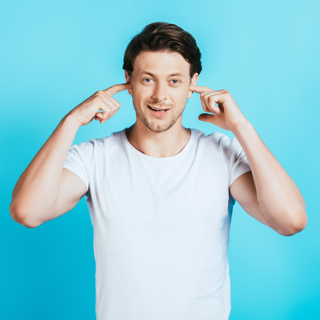 Мужчина в белой футболке закрывает уши пальцами на синем фоне - Фото, изображение
