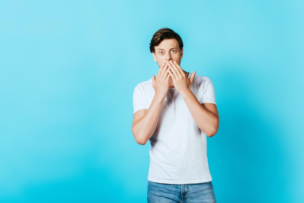 Σοκαρισμένος νεαρός άνδρας με λευκό t-shirt κοιτάζοντας κάμερα σε μπλε φόντο - Φωτογραφία, εικόνα