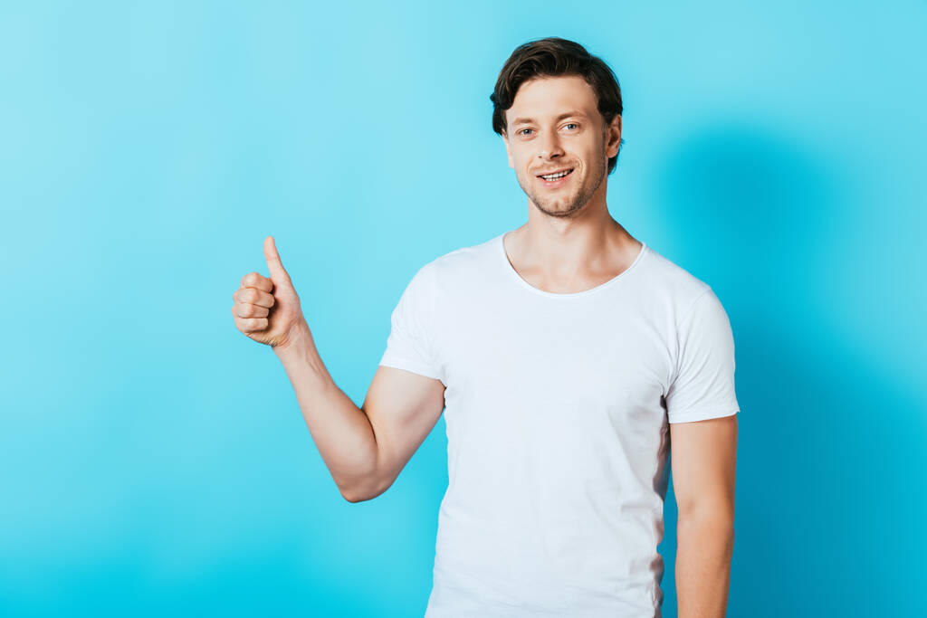 Молодой человек в белой футболке показывает большой палец на синем фоне - Фото, изображение