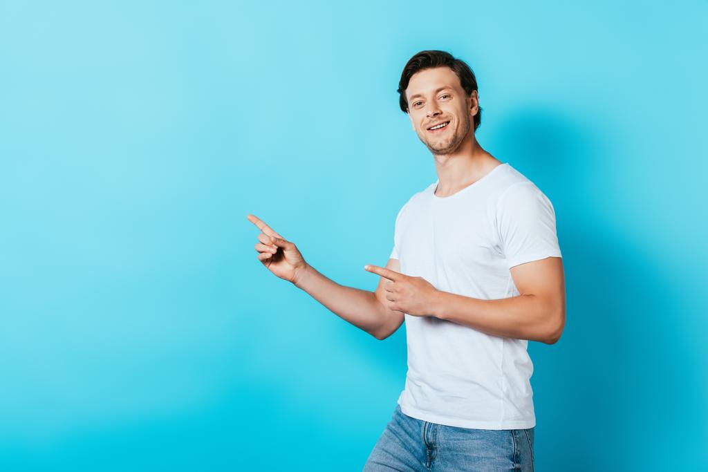 Homme en t-shirt blanc pointant avec les doigts et regardant la caméra sur fond bleu - Photo, image
