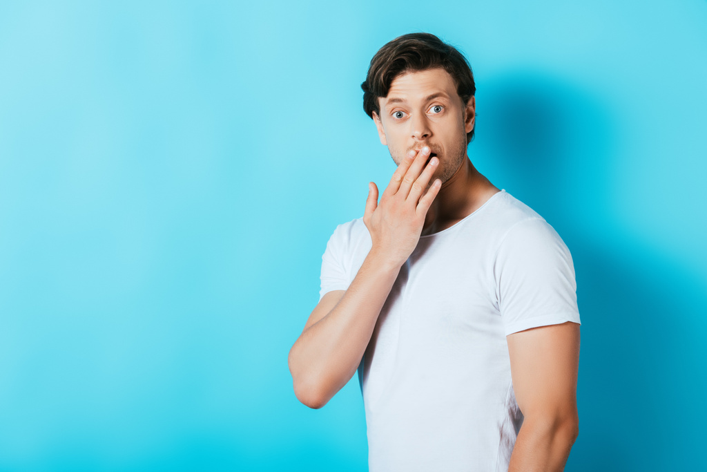 Hombre sorprendido en camiseta blanca que cubre la boca con la mano sobre fondo azul - Foto, imagen