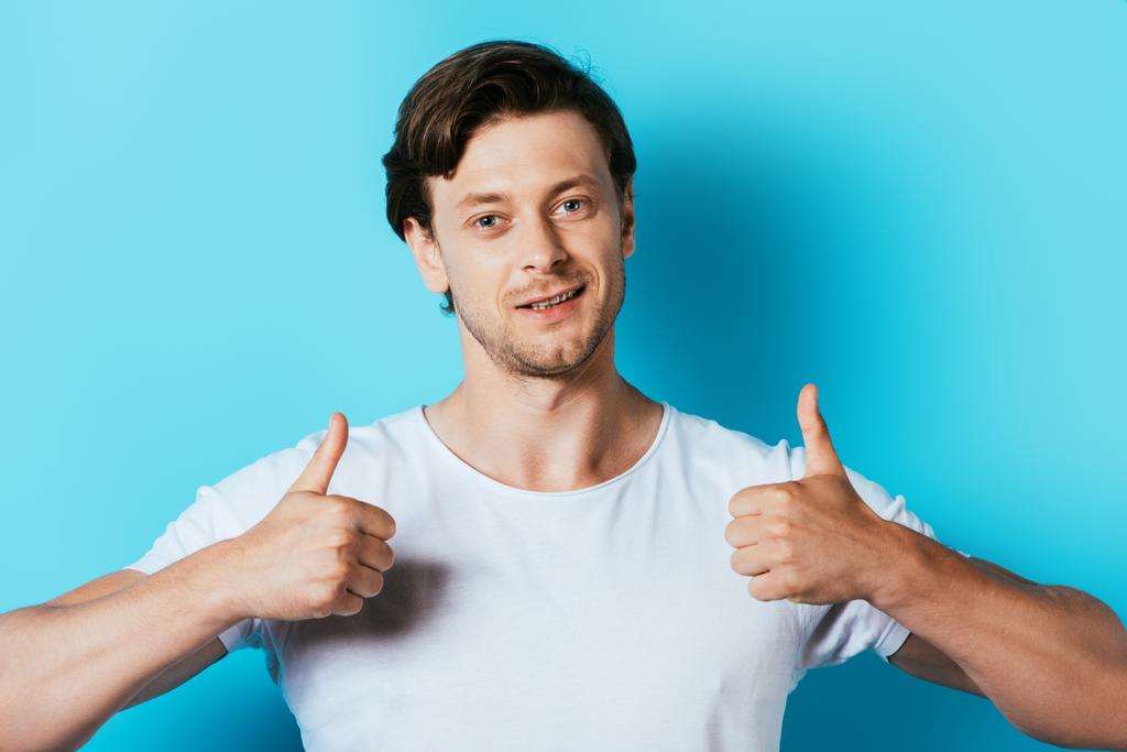 Homem de t-shirt branca mostrando polegares para cima no fundo azul
 - Foto, Imagem